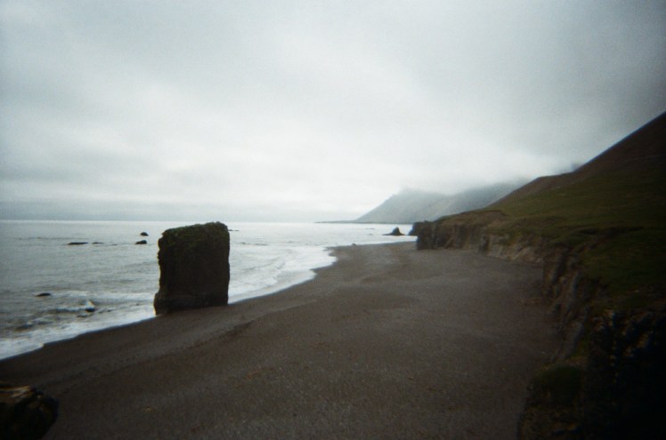 A black beach stretches away into the distance, with a hint of mountains on the right. To the left is a grey sea under grey skies and a rock stack in the middle.