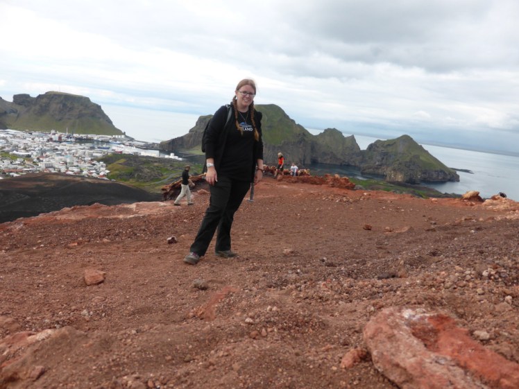 A timer selfie on the top of a volcano. The rock is orangey red and gravelly. Behind me is a steep slope down to town and a zigzaggy cliff on the other side of the harbour.