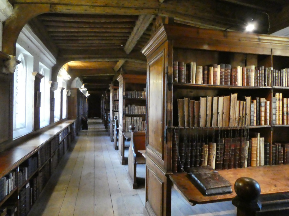 Wells Library. The floor is wooden and the ceiling is made of ancient-looking wooden beams. Under the windows on the left are low bookcases. Then there's a passageway and then rows of tall bookcases perpendicular to the passage on the right. Many of these books look very old and if you look carefully, you can see chains attached to the ones on the middle shelves.