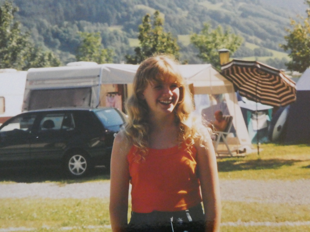 A portrait of me standing in the campsite looking happy. I'm around 13 years old, with long curly blonde hair and wearing an orange strappy top.