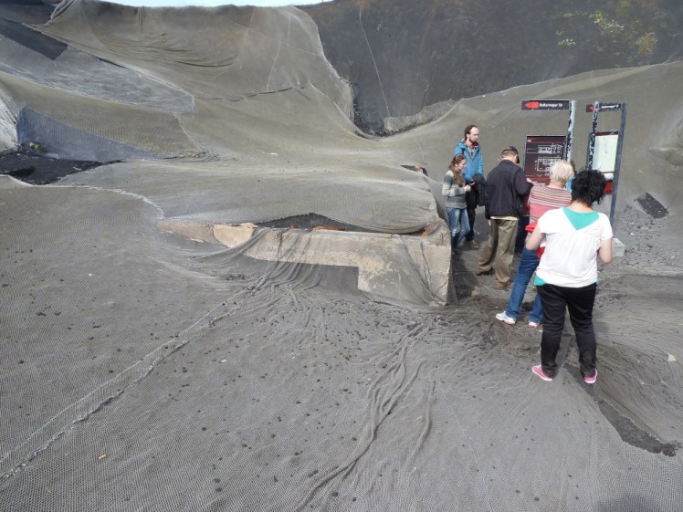 Eldheimar early in its excavation. There's a cluster of people standing next to the corner of a concrete cube which is a house buried up the roof in what looks like grey sand.