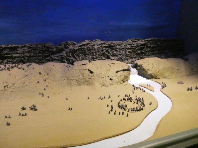 A model of Þingvellir. The sky is very dark blue but the Fields are well-lit. You can see those black cliffs. To the right, a waterfall cascades from near the foot of the cliffs into the fields below. As the river curves round to the left, there is a ring of tiny model people where Parliament would have been, as well as various other people standing on the Law Rock and scattered around the Fields.