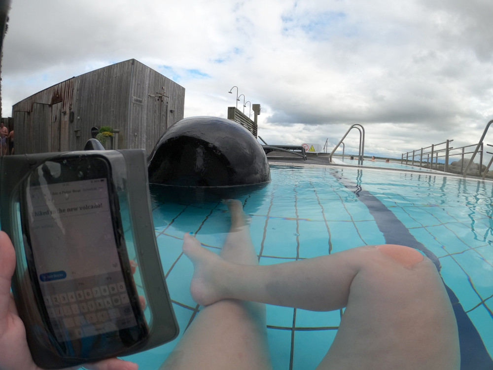 A picture taken from a GoPro (so lots of curvy fisheye edges) of my feet and legs in a shallow blue-tiled pool. In my left hand is my phone in a waterproof case. In front of me is a large black spherical rock sculpture in the water. To the left is a wooden hut containing steam rooms directly over a natural vent, in front are a row of outdoor showers for after your steam room and in front you can just make out a trapezoid miniature swimming pool.