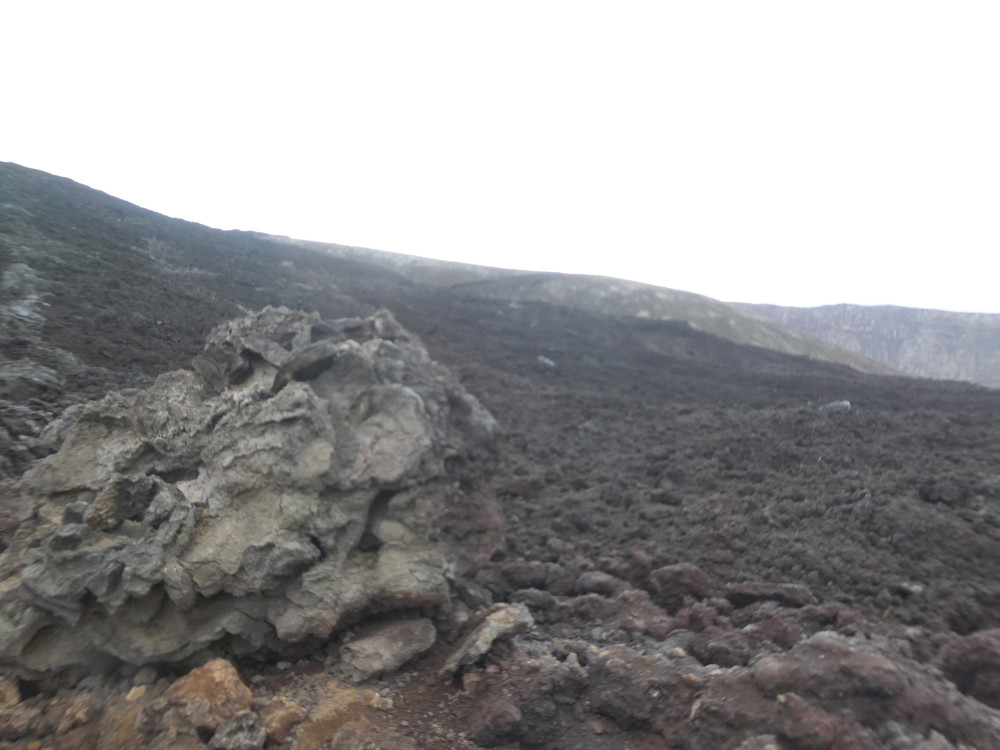 A steep sloping fresh lava field, very rough and dark grey. In the far background are some lighter gentler but higher mountains and in the foreground is a big misshapen rock.