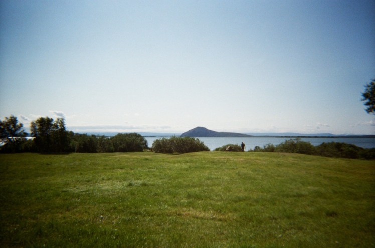 The view across a grassy field and a lake to a mountain, under a blue sky.