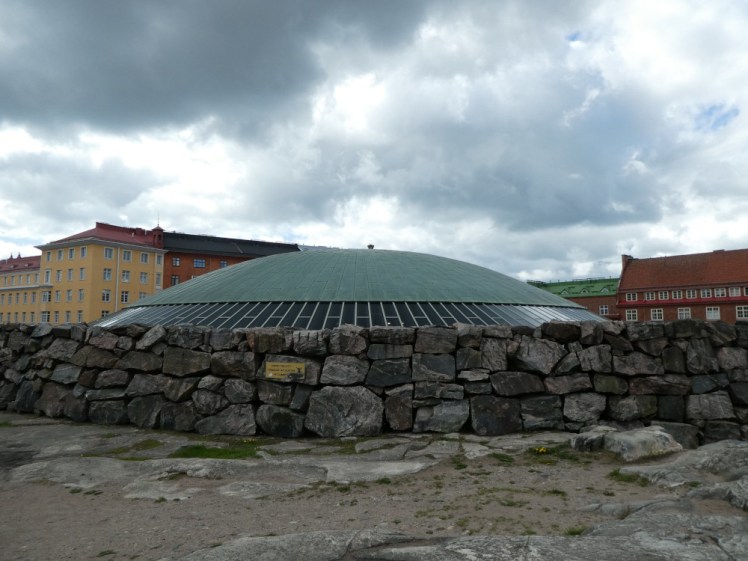 A green copper dome rising out of a low wall of stones. This wall is on top of a hill and surrounded by apartment buildings.