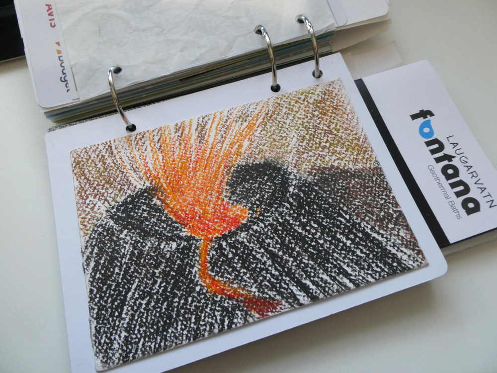 A sketch of the Meradalir eruption in oil pastels, stuck into a small scrapbook bound with three rings. A leaflet for Fontana is sticking out.
