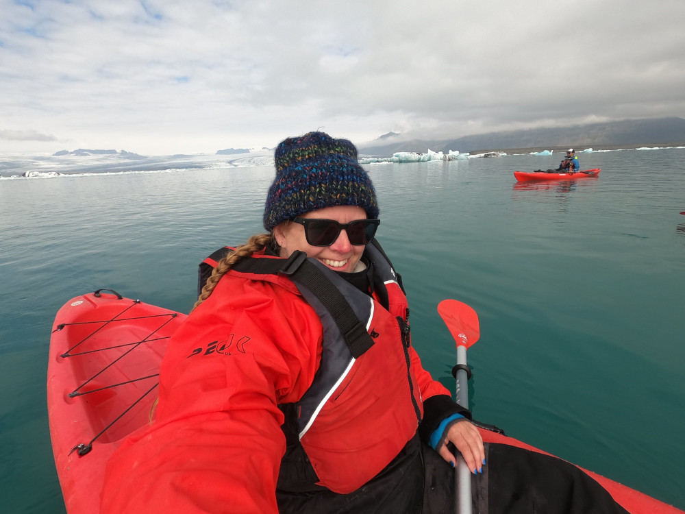 Me, all in red warm & waterproof clothes, sitting in a red kayak on a teal glacier lagoon. The cloud is low, casting some great light on the water.