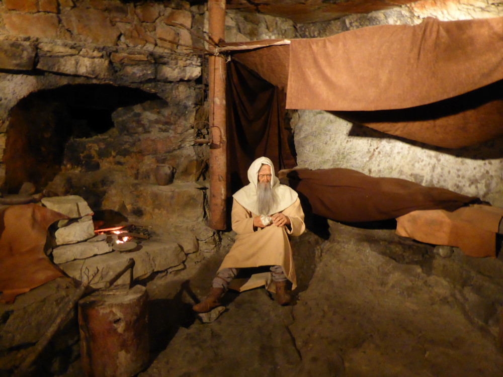 A dummy of St Beatus sitting in a cave with a fireplace to his right and a bed covered in sheets and hangings to his left.