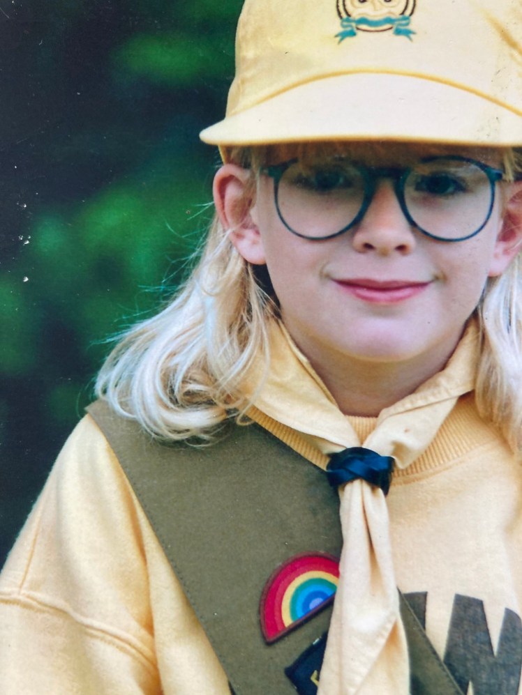 Me as a Brownie. My hair is a lot lighter than it is now, I'm wearing a yellow baseball cap with the Brownie logo on, a yellow sweatshirt with the word BROWNIES across it, a yellow neckerchief held together with a black plaited leather woggle and a brown sash on which you can see my Rainbow badge and the corner of my Six badge.