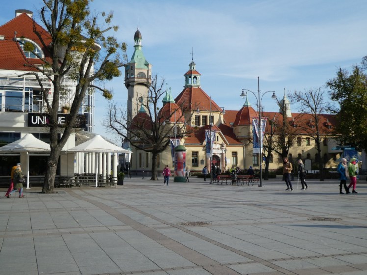 A square in Sopot with an interesting yellow and orange building with a tower along one side of it.
