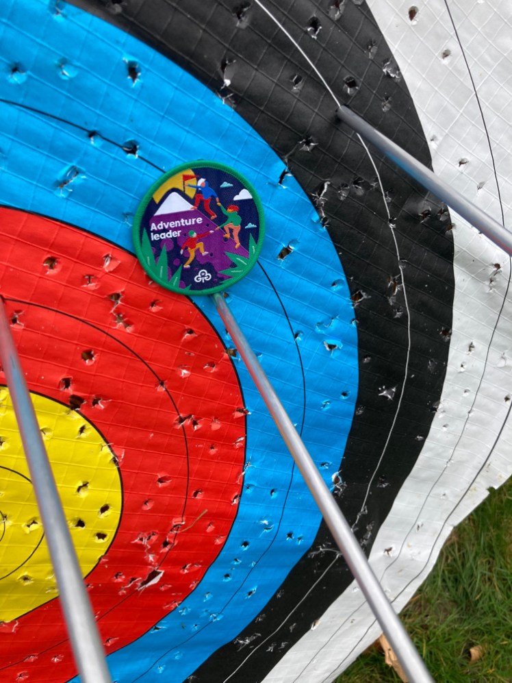 A green-edged "Adventure Leader" badge balanced on an arrow shot into a target. There are two other arrows sticking out of the target.