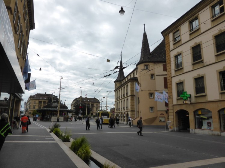 Place Pury, the main square where you usually get the bus, seen from the side of the road leading up to it. There are drainage streams running down the side of the road.