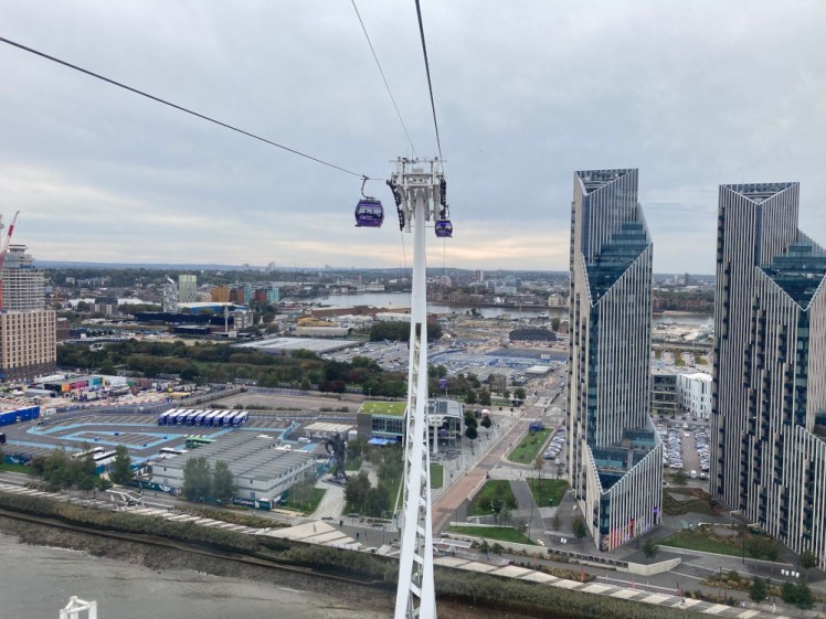 A tall twirling pylon lifts a cable high above east London, almost level with the two shiny tower blocks to the right of the photo. There are two purple cable car cabins hanging from the cable next to the pylon.