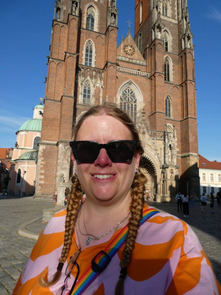 A selfie in front of the tall brick Gothic facade of the cathedral. I'm wearing sunglasses, a pink and orange t-shirt and I look pink and sweaty.