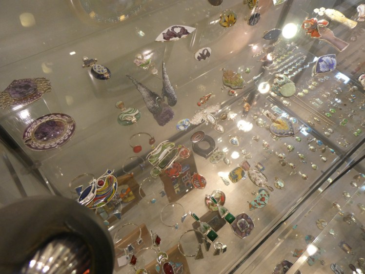 Looking down through glass shelves of enamel jewellery, ranging from small things like earrings to large brooches.