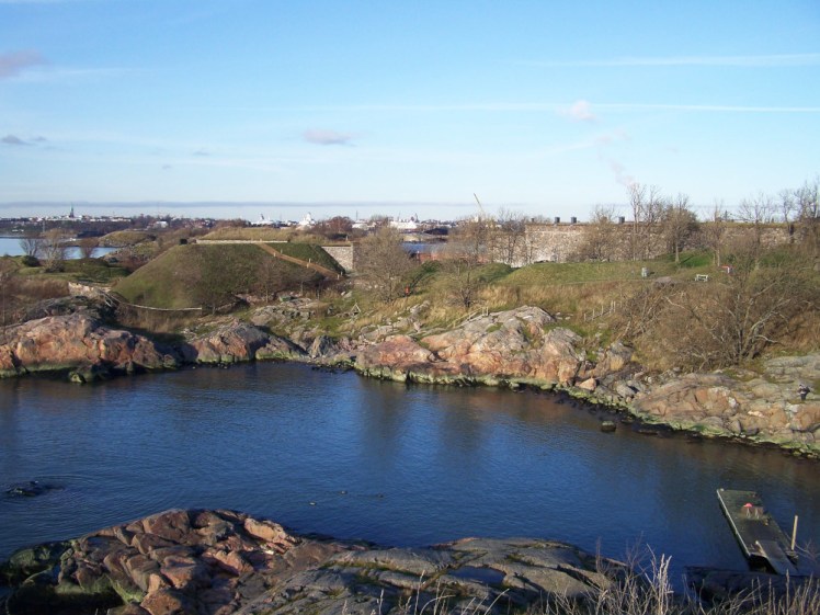 A view from Suomenlinna. In the foreground is a small dark blue bay surrounded by tiny cliffs. On the other side is a flat-topped hill that's probably a fort, and there are various ex-military buildings around. Above and behind them, you can make out the skyline of Helsinki.