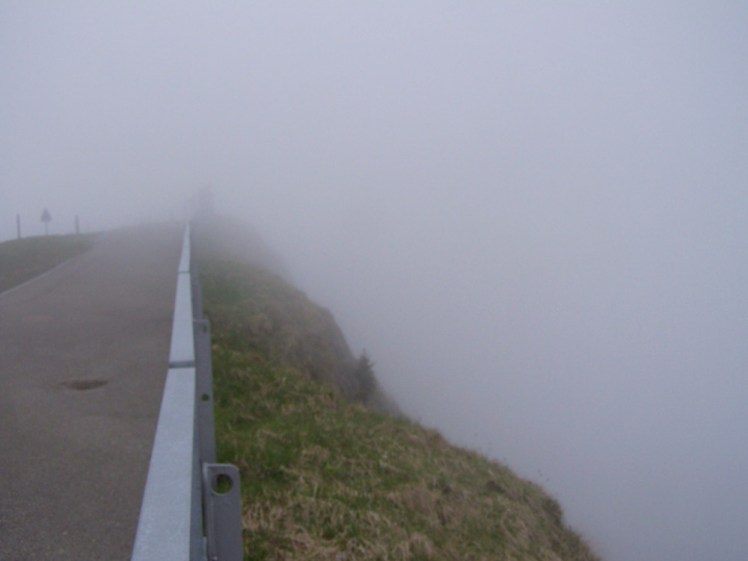 A grey crash barrier separates a concrete path from the grassy side of the mountain. The mountain drops sheer away but you can't see anything because the rest of the picture is enshrouded in the sort of thick grey fog that obscures everything including daylight.