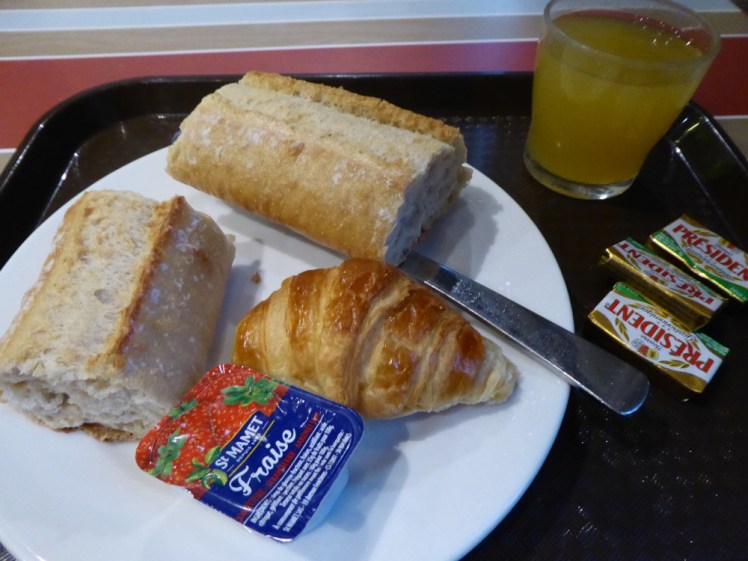 Hotel breakfast in an Ibis in Paris: A white plate on a black tray. On it: a glass of orange juice, three mini packs of butter, two pieces of crusty baguette, a small croissant and a sachet of strawberry jam.