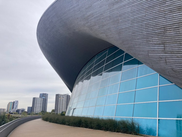 A massive curved blue glass window at apparent ground level (the pool is actually at least one storey down) with a big curving "browbone" of a roof swooping up and over it. This is the side and the roof of the pool building.