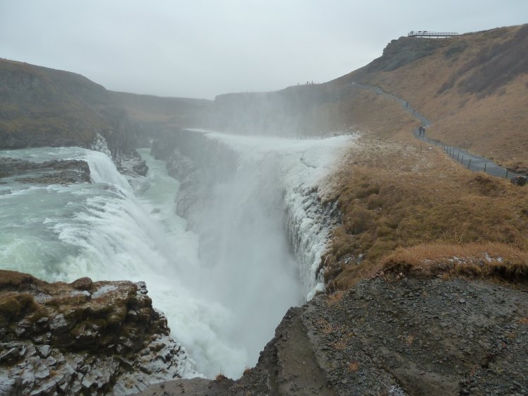 Gullfoss, taken from the rock next to its second cascade. The grassy banks are orange and deep yellowish green because it's autumn. The water is a light teal green, partly frozen already, and is falling into the chasm below and sending up a lot of spray, even at less than full power.