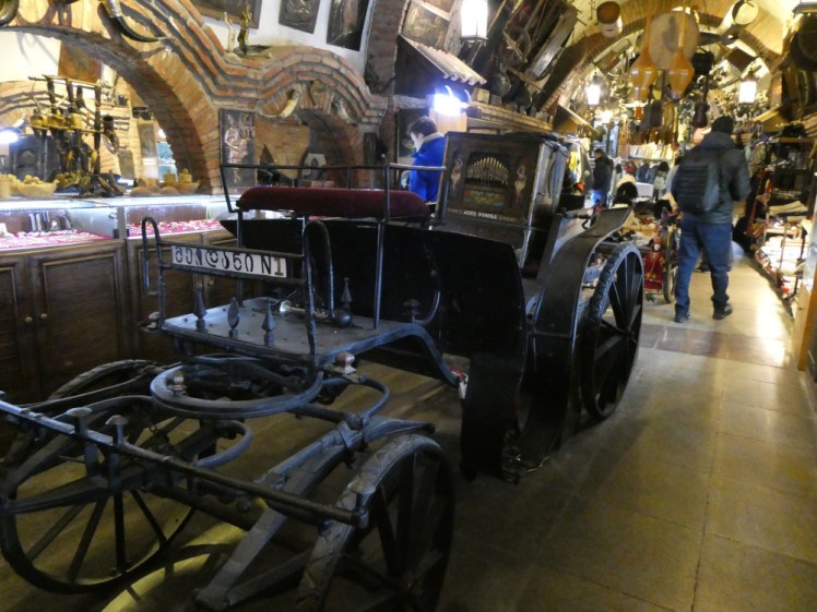 An old, probably horse-drawn car, inside the bazaar. Cupboards along the side of the passage have glass display cases above and there's stuff hanging from the higher walls and the ceiling all the way along.