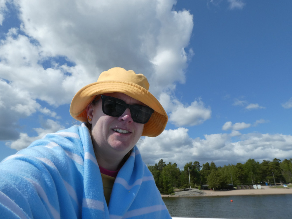 A selfie wearing a yellow bucket hat, sunglasses and a pale blue blanket because it's freezing. I'm on a boat and behind me is a bit of water and then one of Helsinki's many forested islands.