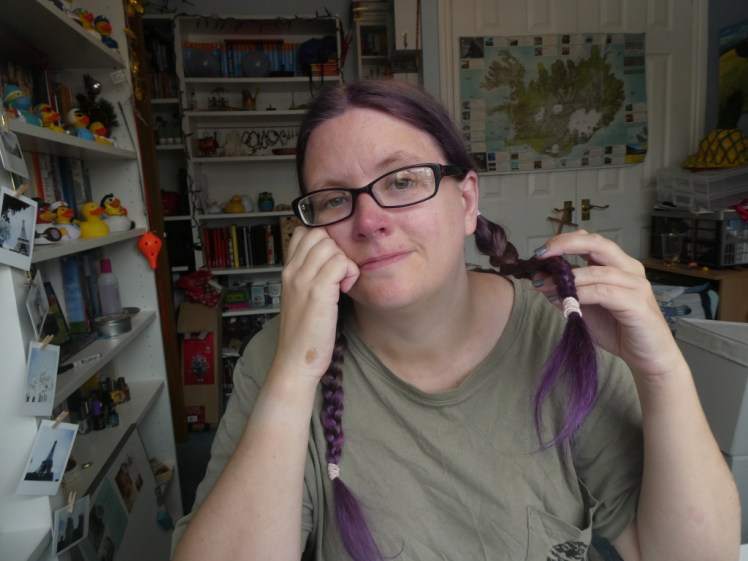 Me, sitting at my desk. My hair is purple and in two plaits, I'm wearing a greyish-green t0shirt
