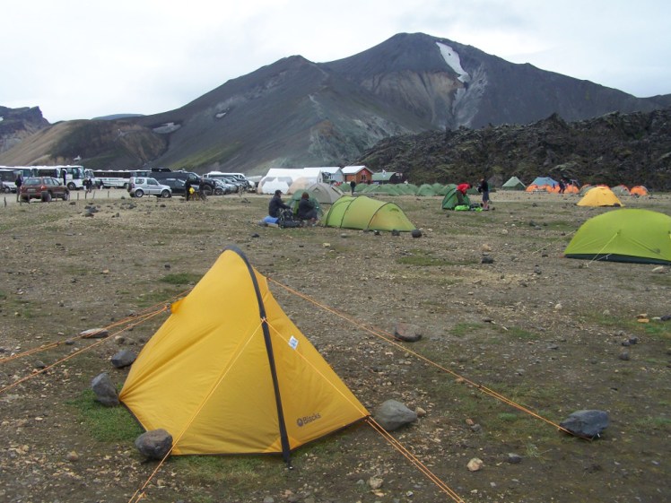 Camping in windy conditions at Landmannalaugar. My tent is a tiny one-man yellow one, weighed down by rocks, on a campsite that looks like bare rock and overlooked by a dark looming mountain with a patch of snow on the top.