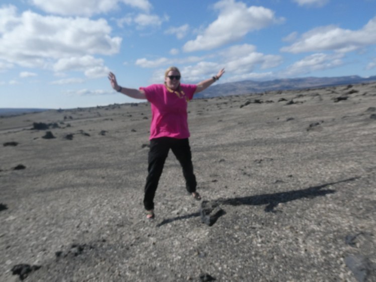 My "moon photo", a blurry selfie taken using the timer functin. I'm wearing a big pink t-shirt and am in mid starjump. Behind me, the land is grey and bare with only a mountain on the horizon.