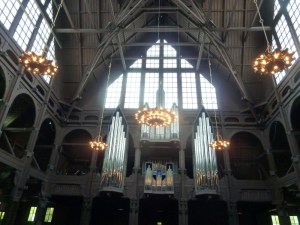 The interior of Kiruna Church. It's all dark wood again with a kind of scaffolding-like sculpture. There's an organ high on the wall opposite and a large non-stained glass window above. There are orange-bulbed chandeliers hanging down.