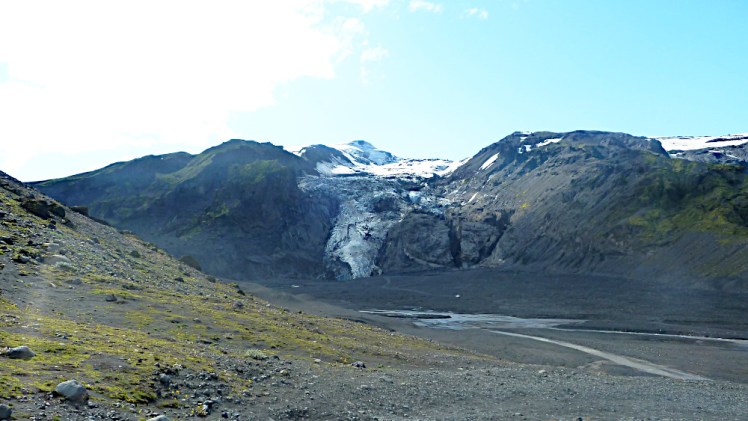 A glacier falls abruptly off the side of a mountain into a gravelly bowl that once contained a lagoon with chunks of ice.