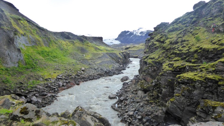 A small river bubbling through a shallow-sided canyon. Around the river there are lots of boulders, the slopes are mossy and in the background is a snowy mountain.