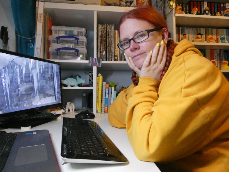 Me sitting at my desk back when the shelves were less cluttered. I have red hair in plaits and I'm wearing a yellow hoodie with matching neon yellow nails.