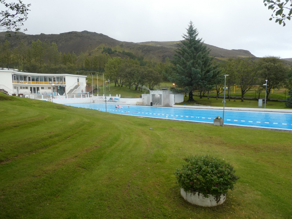 The public pool at Hveragerði, a slither of bright blue on the side of a green hill with a mountain above it.