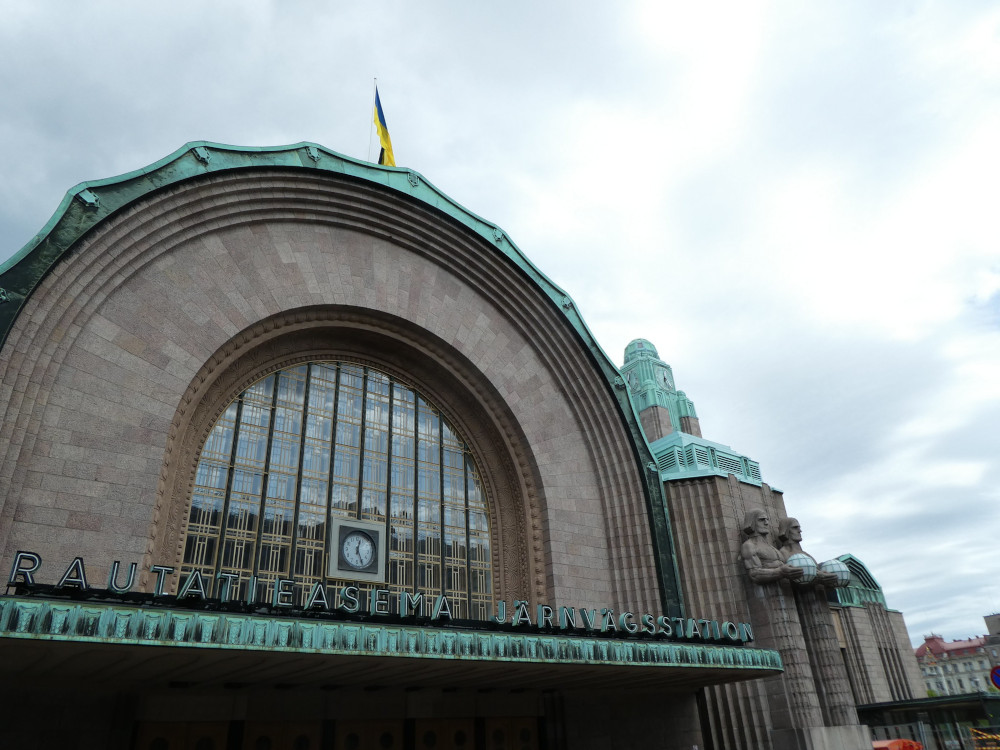 Helsinki Central Station, a building of pinkish-brown brick with copper-green accents, like a frill around the great arched front and along the front. There is a green-topped tower just visible. The doors are flanked by two giant figures on each side holding a glowing green globe. It's built in the Modernist style.