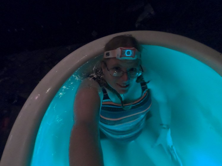 Me in the hot tub after dark with the camera held above my head. I'm wearing a striped swimsuit and a headtorch and the water is lit up in light blue.