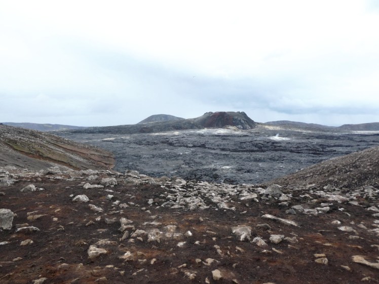 The Fagradalsfjall craters in a sea of grey-black lava in the distance. The photo is taken from a viewpoint that's made of older, browner lava with a thin layer of soil overlaid.