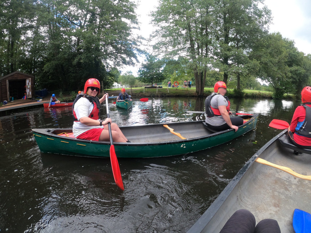 Me in the back seat of a green two-person canoe. I'm wearing my grey t-shirt and red shorts and my faceless partner is wearing a grey t-shirt. Behind us are a few more canoes and there are people on the bank behind us who I think are pioneering rather than watching us.