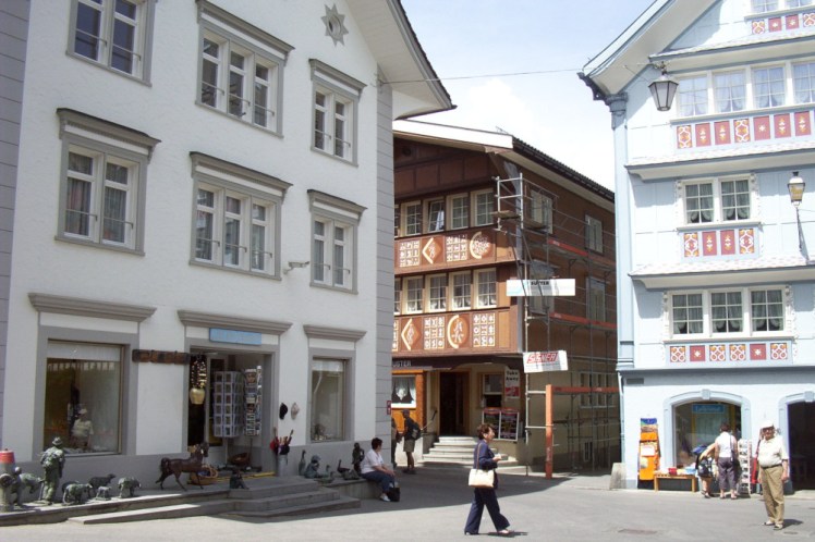 Appenzell village centre, three three-storey chalets, two painted white, one with balconies across the front, next to a brown wooden one, also with balconies. The balconies on both are painted with red and yellow decorative motifs.