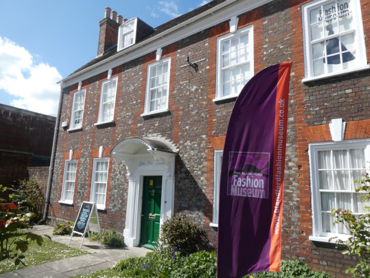 The Blandford Fashion Museum, a brick Georgian house with a purple and orange museum flag in the front garden. (This isn't actually the main door but it's the prettier side of the place).