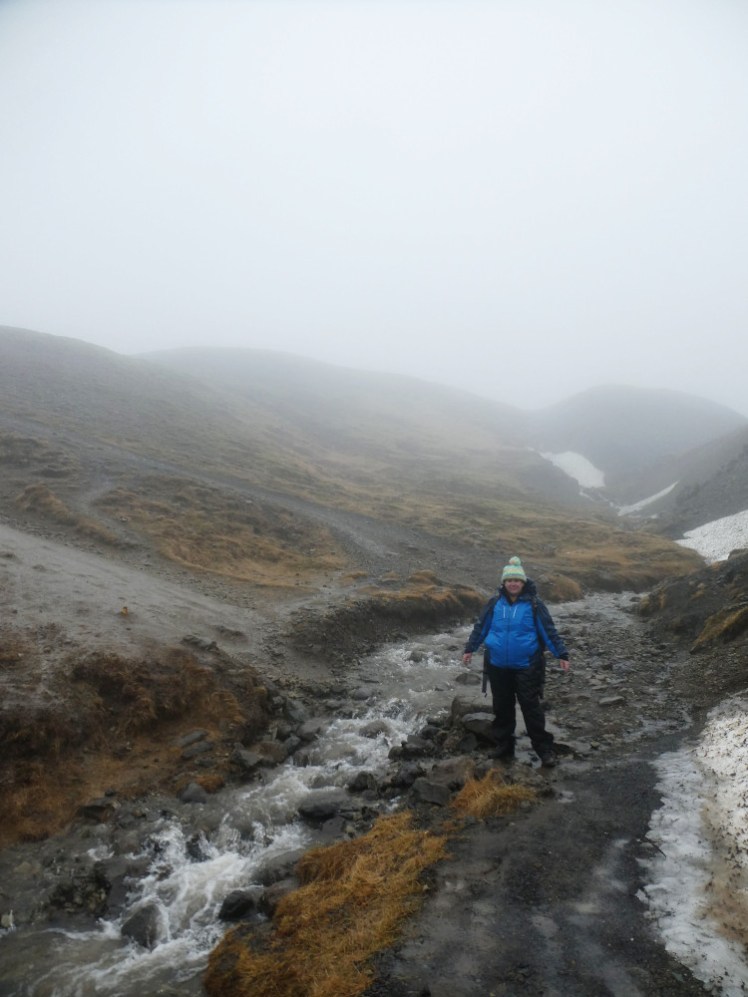 Me, in my blue raincoat and rainbow-white hat, standing next to a small bubbling river. The path is just black mud, the mountain behind me is grey, orange and khaki streaked and fades into mist in the distance.