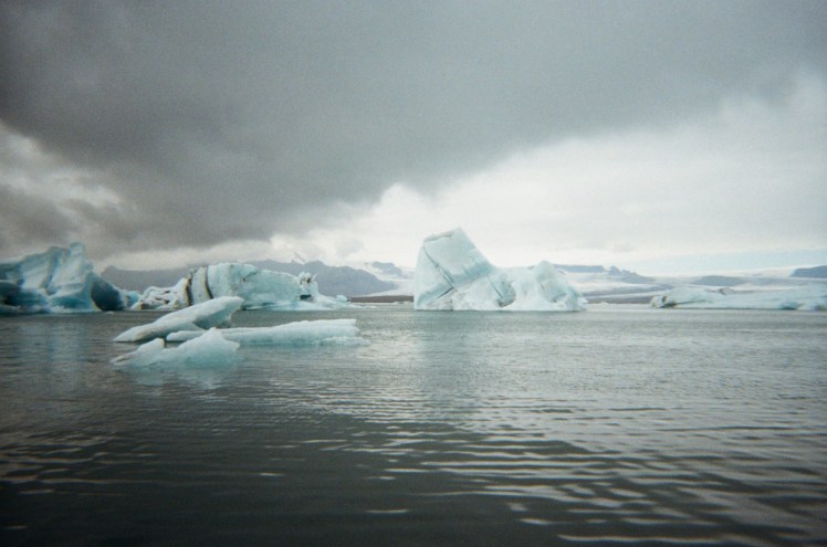 A 35mm film photo of icebergs floating in Jökulsárlón. There are big heavy dark grey clouds overhead. The grainy film colours really add to the atmosphere of the glacier lagoon.