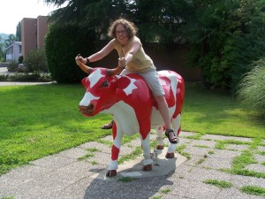 Angela sitting on the plastic/fibreglass red cow outside the factory. She's leaning forward to hold onto its horns.