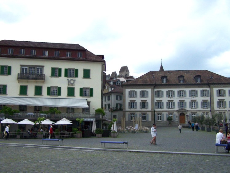 My hotel in Zurich, a pleasant-looking stone building with green shutters and seats under umbrellas outside in a cobbled square.