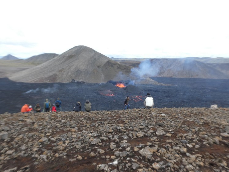 The 2022 eruption when it was still live - a tiny black crater filled with exploding orange lava, in what was once a valley, now filled with fresh black lava flow. Behind it is a larger greyer gravelly hill and there are more gravelly grey hills behind containing the lava flow. The photo is taken high up on a rocky hill overlooking the eruption. There are lots of people sitting and standing on the hillside watching - and a lot more off to the sides and further down the hill than you can see from this angle.