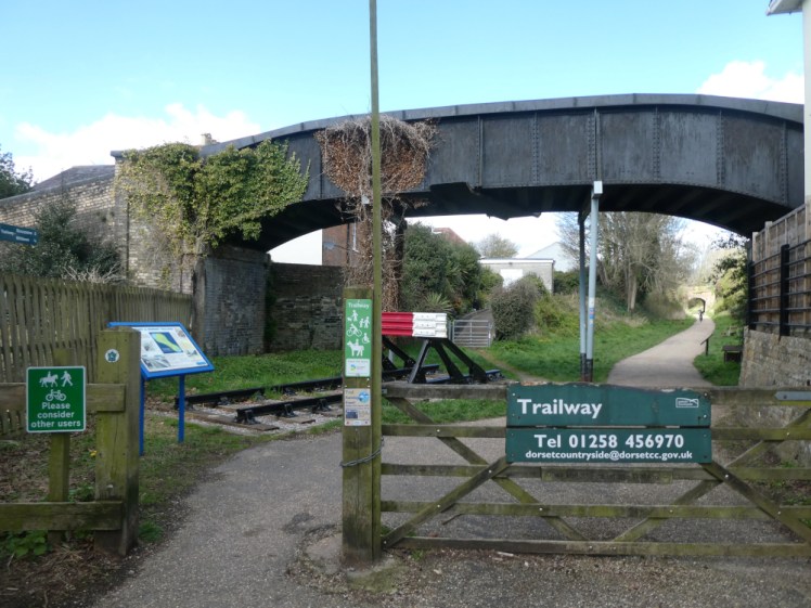 The Blandford end of the North Dorset Trailway. An open gate leading onto a gravel track between steep grassy banks. A thin railway footbridge runs over the top.