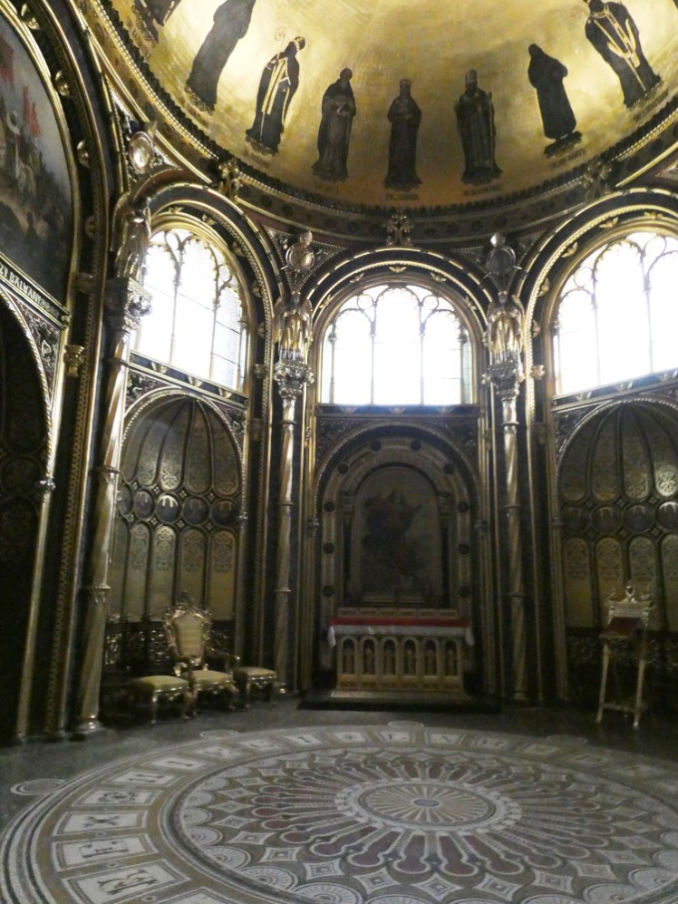 The Golden Chapel. It has golden walls and a domed ceiling and rounded arches and overlit stained glass windows.