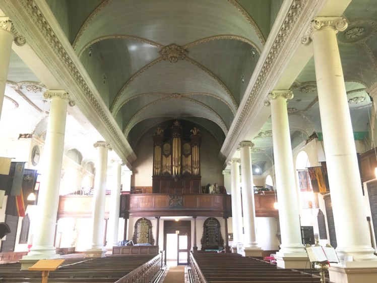 Inside Blandford's church, an over-lit picture of a church with dark wooden pews and organ but white pillars and supports.