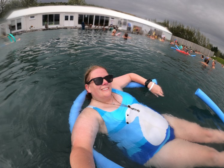 Me in the Secret Lagoon, floating on two blue noodles. I'm wearing a one-piece swimsuit striped in various shades of blue underneath a large polar bear sitting down looking over its shoulder.